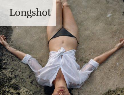 From above of full length slim female with closed eyes in white shirt and black bikini lying on sandy shore in water while enjoying time