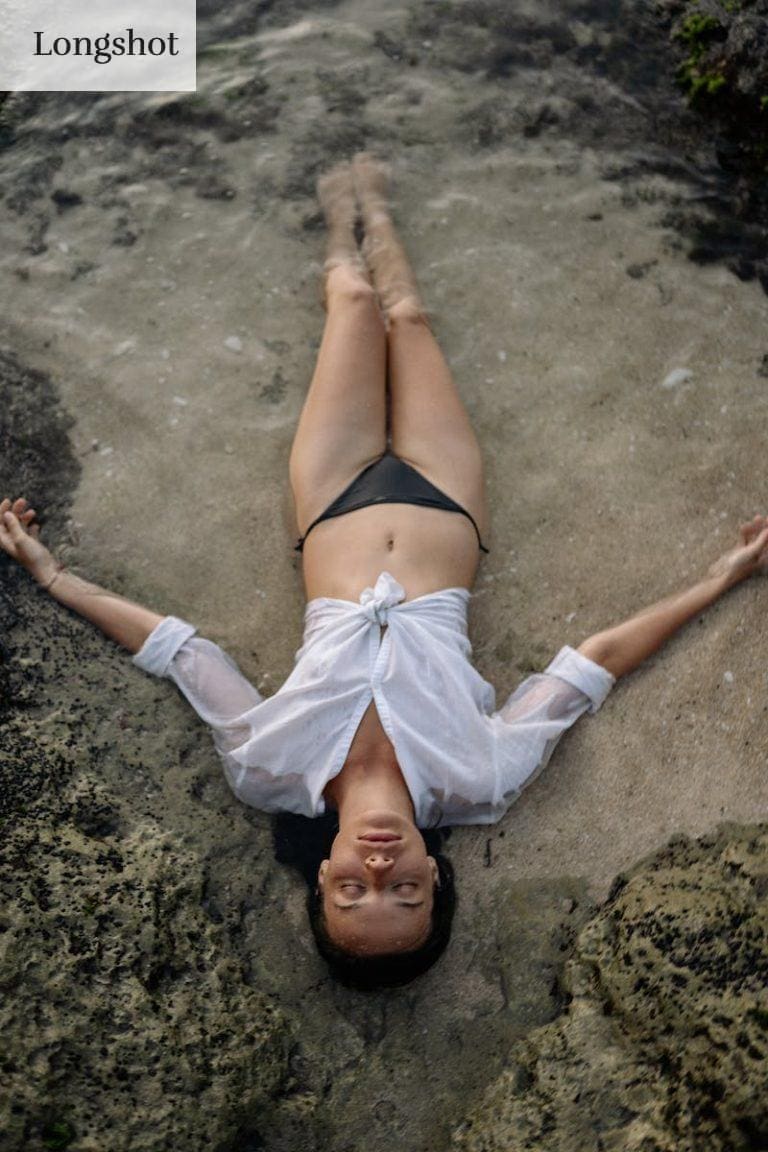 From above of full length slim female with closed eyes in white shirt and black bikini lying on sandy shore in water while enjoying time