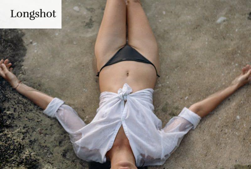 From above of full length slim female with closed eyes in white shirt and black bikini lying on sandy shore in water while enjoying time