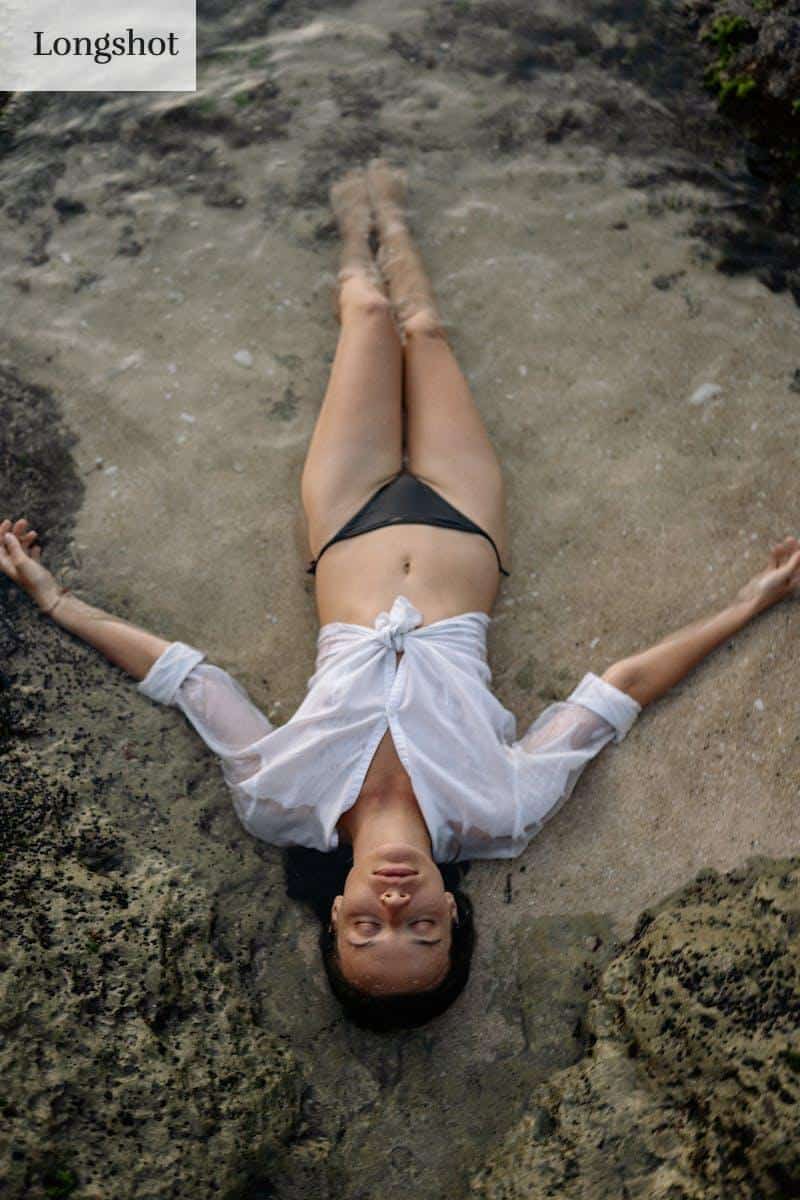 From above of full length slim female with closed eyes in white shirt and black bikini lying on sandy shore in water while enjoying time